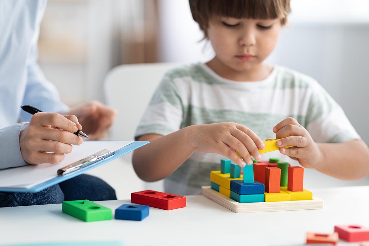 Close up portrait of cute little boy during play therapy at child psychologist's office, professional woman with notepad making notes