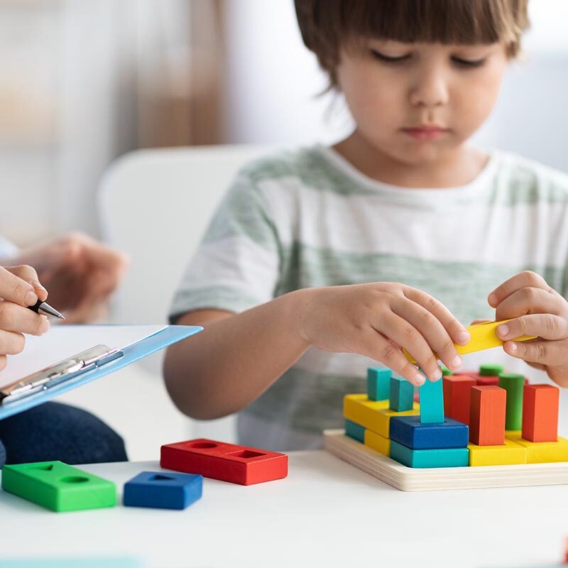 Close up portrait of cute little boy during play therapy at child psychologist's office, professional woman with notepad making notes